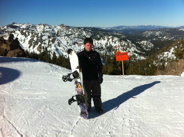 A man poses with a snowboard at the top of a mountain covered in snow. An orange sign warns of a hard no-pass zone.