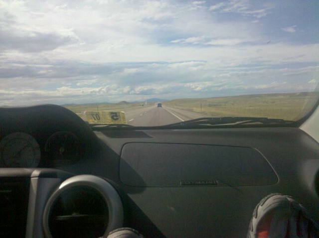 The dashboard of a car, looking out at a long road in middle America.