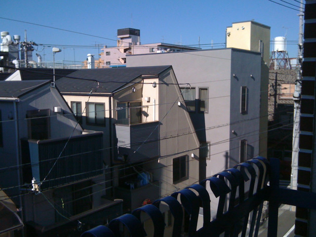 A view from a window into a Tokyo neighborhood. The building across the street has some laundry drying. The sky is blue and clear.