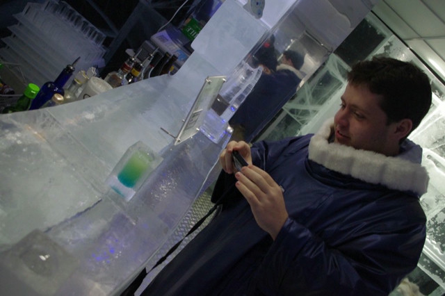 A man takes a photo of a drink in a bar fully made of ice. He's wearing a parka due to the temperatures required to keep the bar from melting.