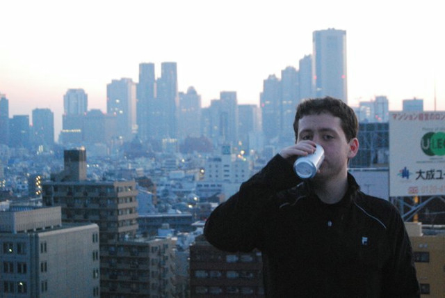 A man sips a beer while the sun rises over Tokyo in the background.