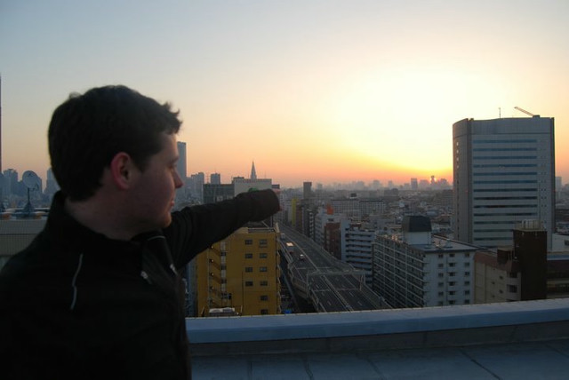 A man in a black jacket pointing at the sun rising on the horizon.