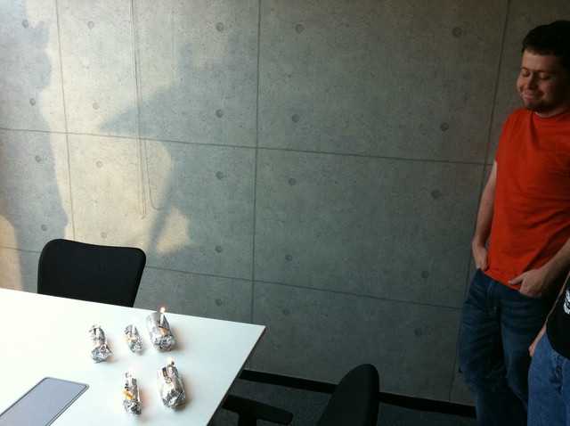 A man in jeans and an orange shirt smiles. There are burritos on a table in front of him.