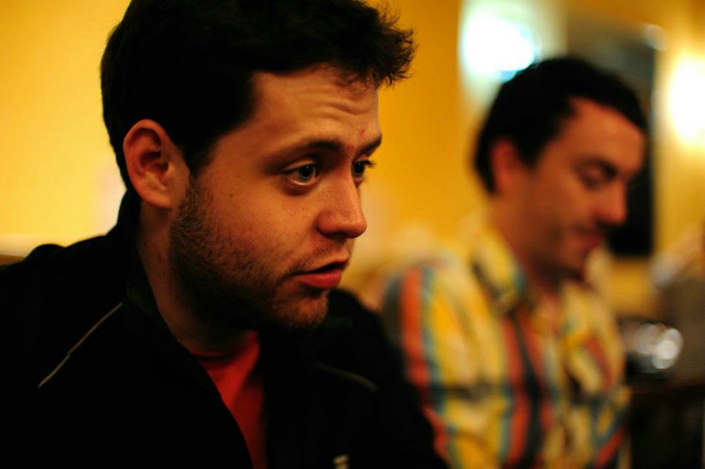 A man with dark circles under his eyes, wearing a black track jacket. In the blurred background is another man in a plaid shirt.