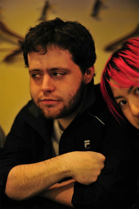 A man with dark circles under his eyes, wearing a black track jacket. A woman with pink hair pokes her head in from the right side.