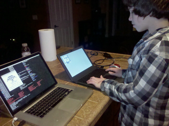 A man stands at a counter with two laptops, each running a compile job.