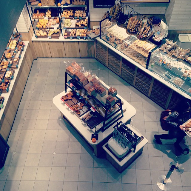     A top-down view of a bakery. Baked goods line the walls, while a worker
    restocks items and a customer finishes browsing.
