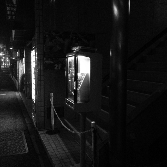 A dark alleyway, with a bright Japanese phone booth centered in the shot.