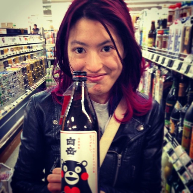 A purple haired woman in a leather jacket holding up a bottle of alcohol.