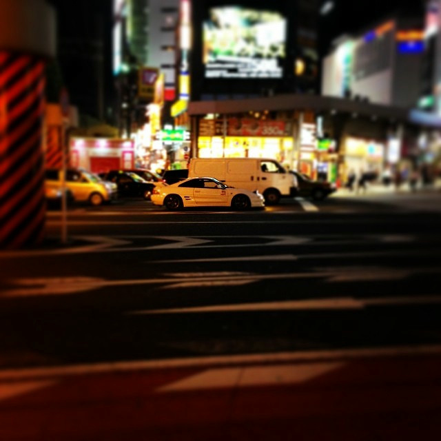 A white Toyota MR2 sits in center frame, amidst a backdrop of traffic and people.