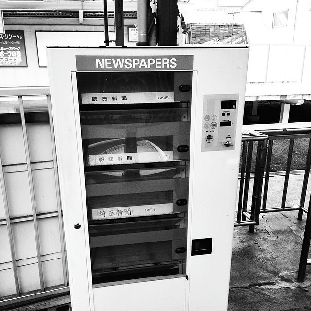 A square Japanese newspaper vending machine on a train platform.