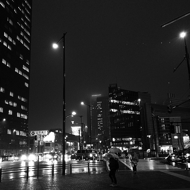 A rainy Tokyo intersection, black and white. Traffic is heavy and umbrellas are out.