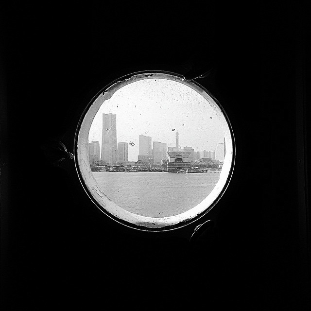 A dark photo with a ship porthole window in the center, looing at the shore. Buildings are on the shore.