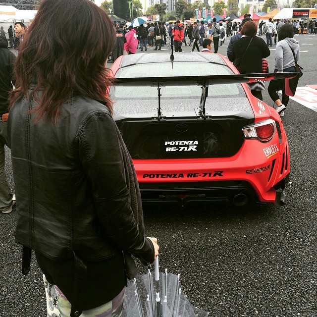 A woman stands behind a car. The car is red with black accents. The woman is wearing a leather jacket and has dark red hair.