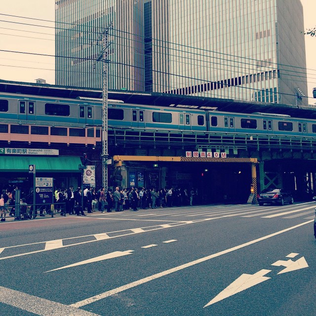 A long line under a train tunnel. A train is picking up passengers above.