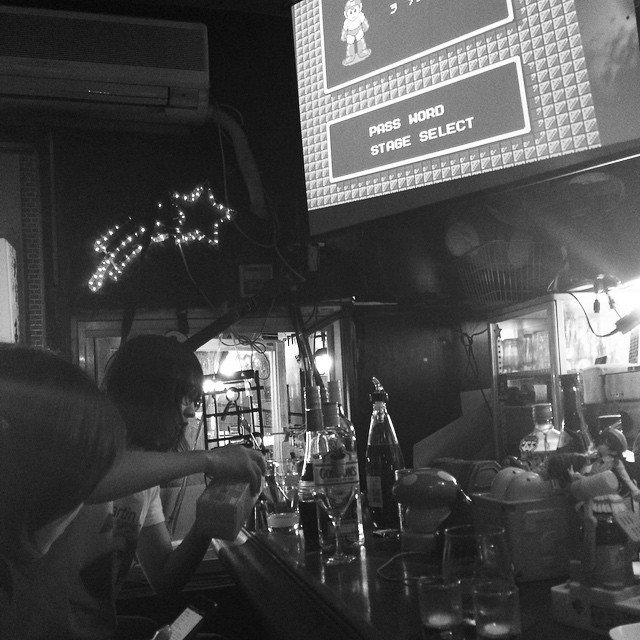 A bartender pours a mixed drink while a customer looks on. Above is a projector that's display a cutscene from a videogame.