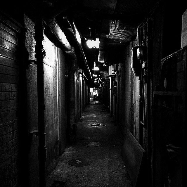 Looking down a very dark and dimly lit alleyway somewhere in Tokyo. Pipes line the walls and are barely visible. At the very end, a sign for a bar is barely visible.