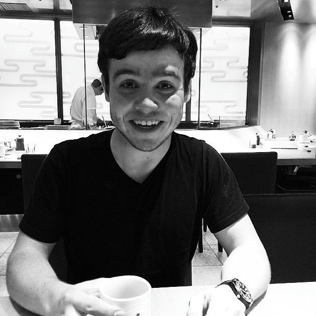 A black and white photo of a man smiling in a restaurant. He's wearing a watch and holding a small cup of tea. A chef works diligently in the background.