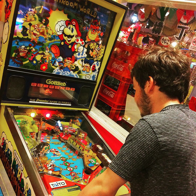 A man hunched over a Mario-themed pinball machine. The machine is yellow with red and blue accents.