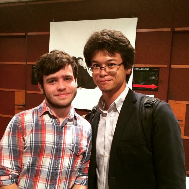 Two men stand next to each other. They are both named Nick. A sign in the background points to the media lounge.