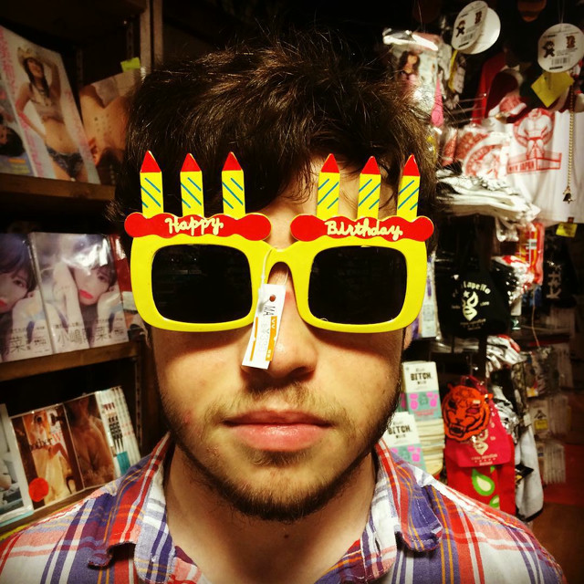 A man poses in a store wearing a pair of glasses that say 'Happy Birthday'. In the backround is a variety of illicit apparel.