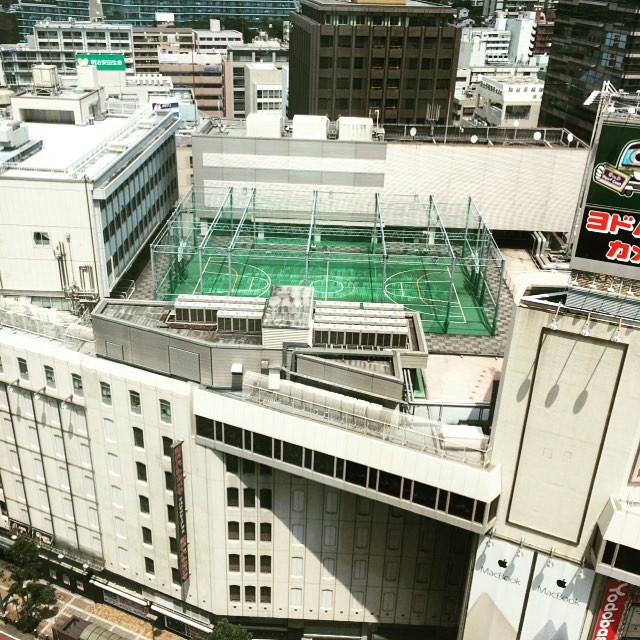 A boring white department store with a hidden green basketball court on top. The basketball court is inside of a chain link fence.