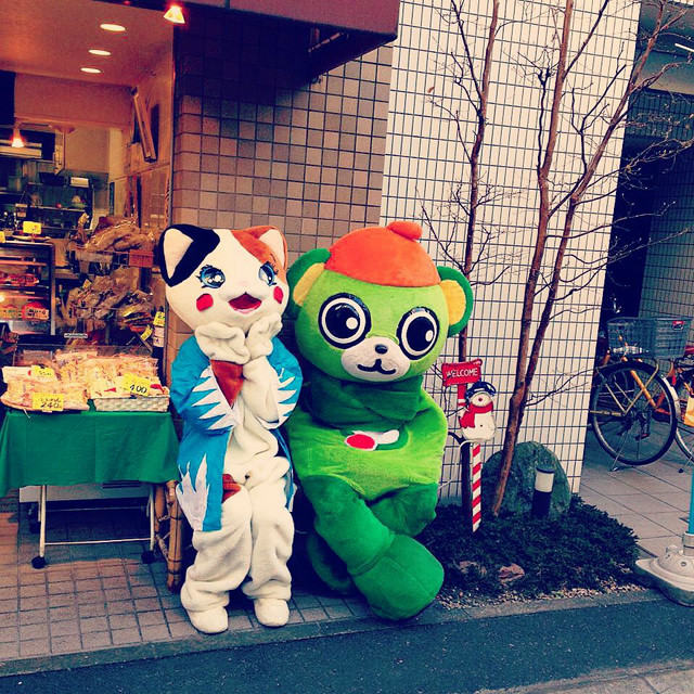 Two mascots sit on a bench. The green bear mascot has arms folded, the white cat mascot is making an excited face.