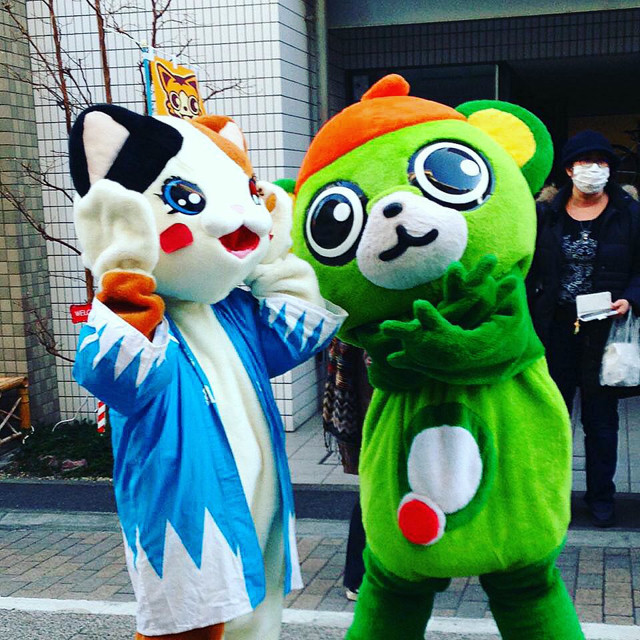 Two mascots stand in a shotengai (shopping street). The green bear mascot has arms folded making a sign, the white cat mascot is making an excited face.