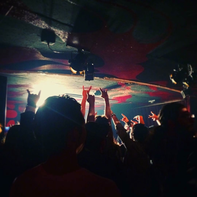 A crowd throwing metal horns in the area as the show ends. The venue has blue and red walls, slightly lit up by the stage lights.