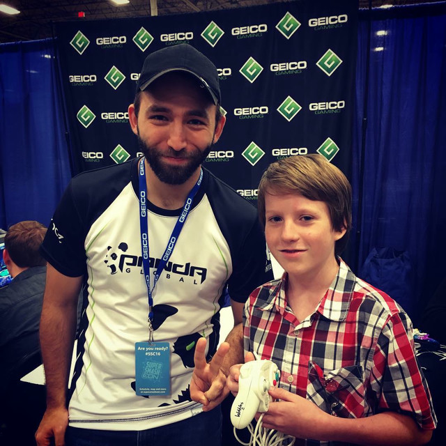 A boy holds a Gamecube controller signed by the player 'Wobbles', who's standing next to him. Geico logos from a sponsorship in the background.