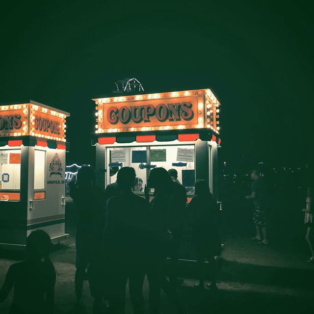 A lit-up coupons stand for a carnival at night in the middle of a field.