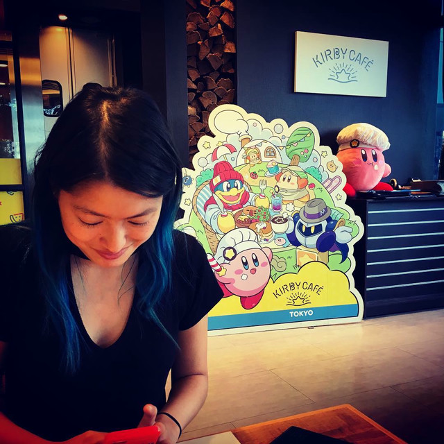 A woman looks down at her phone in a cafe. The cafe is kirby themed. The sign for the cafe is behind the woman.