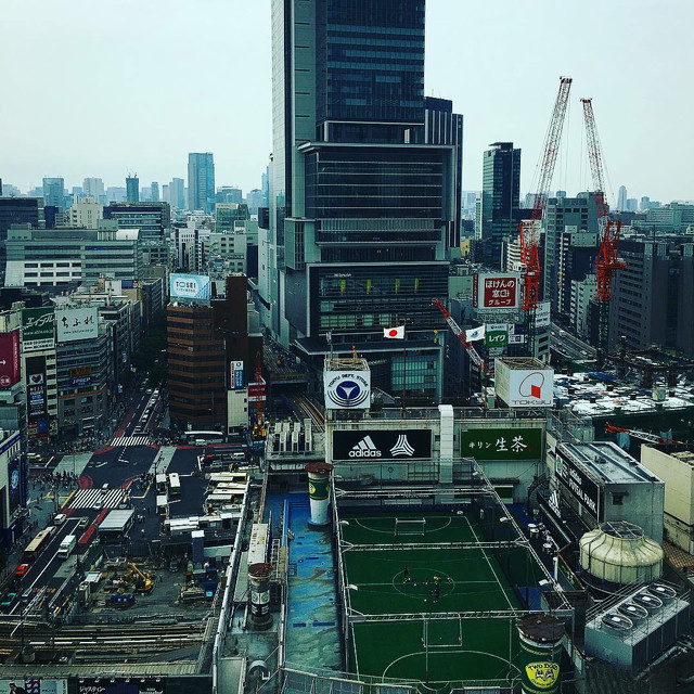 A wide shot of the Shibuya area. A bus terminal is visible, with buses on the move. A soccer field sits atop a building, with cranes everywhere.