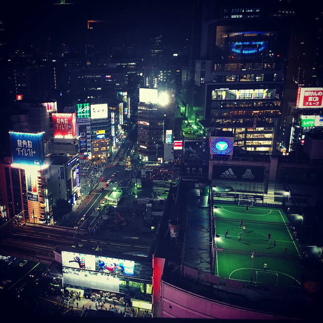 Shibuya lit up at night. People are playing soccer in a field on a roof.