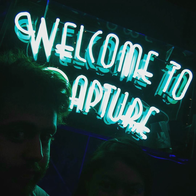 A selfie of us in front of a neon blue sign that says 'Welcome to Rapture'.