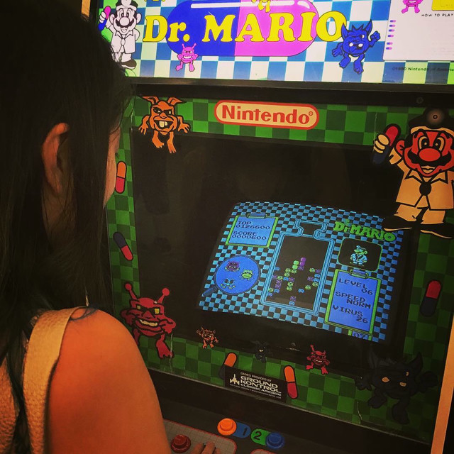 A woman stands over a green Doctor Mario arcade unit. She's on level 6. The unit is sourced from Ground Kontrol in Portland, Oregon.