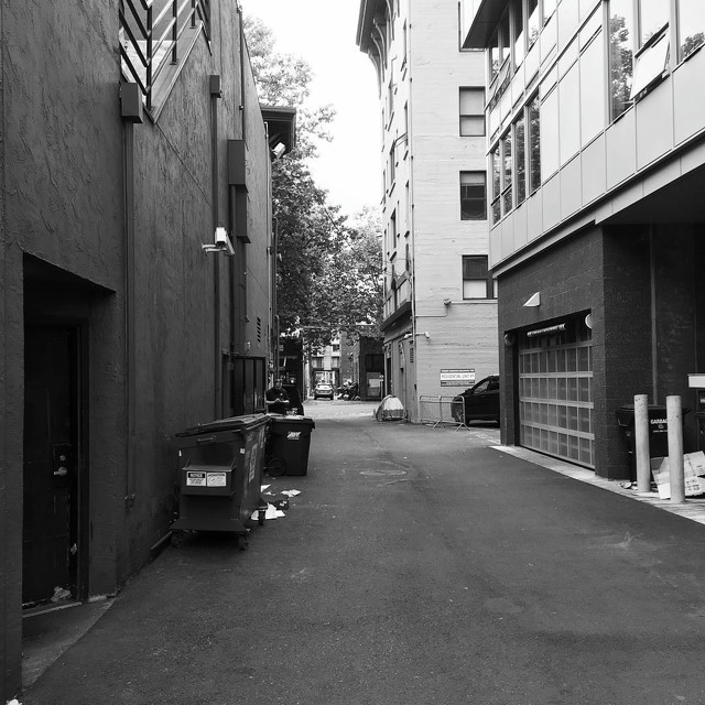 A view down an alleyway in Belltown, Seattle. There's a dumpster on the left and a garage door on the right. At the end of the alley is a cross street with cars waiting.