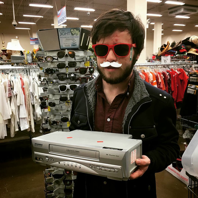 A man with fake glasses and a mustache holds a VCR in a store.