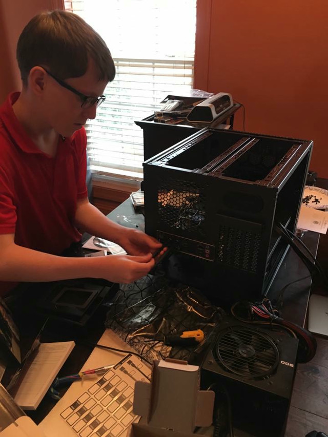 A boy in a red shirt is screwing together a black computer case on top of a wood desk.