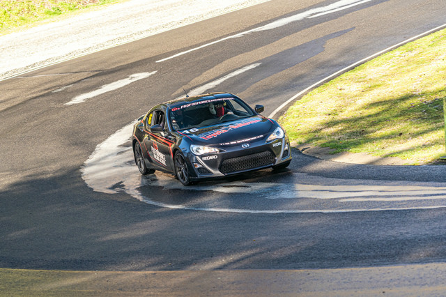 Myself in a black coupe, going through a left hand downhill turn. Myself and my exam instructor both are wearing masks obscuring our faces, unfortunately. The road is slightly wet, with rain shimmering on it.