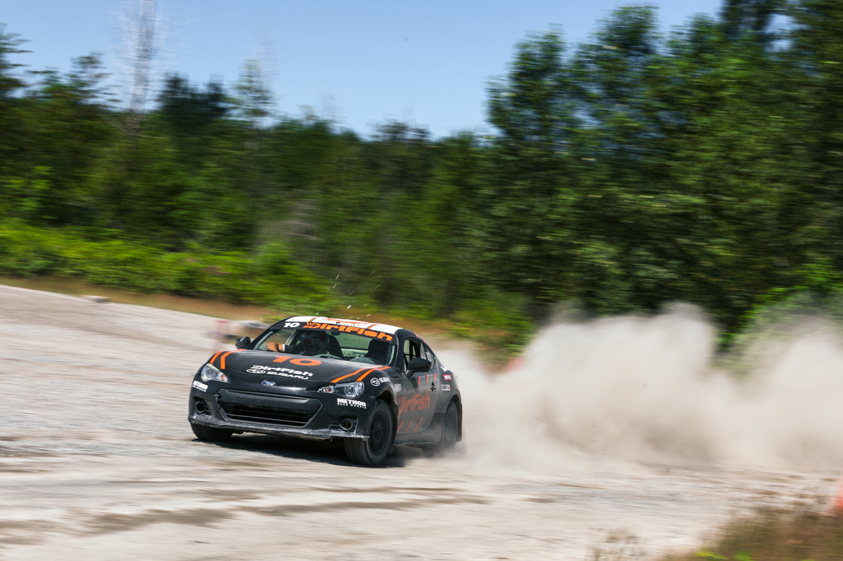 A black car with orange highlights drifts out of a wooded area. Dust clouds are following.