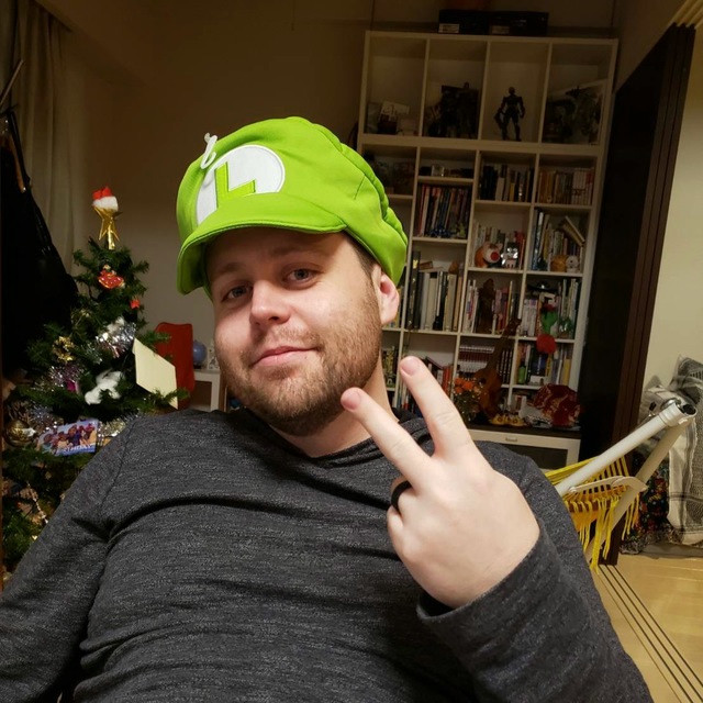 A man poses in a green Luigi hat from the Mario Bros videogame series. He's also making a peace sign.