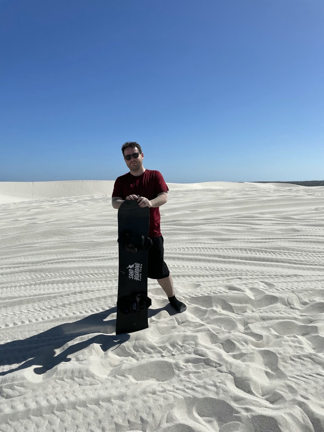 Me posing on a white sand beach dune with a sandboard.