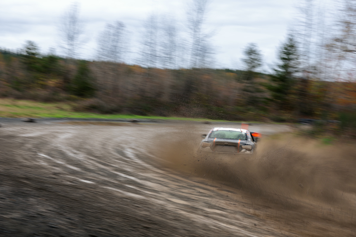 A black car with orange highlights, sliding through a right hand turn. The turn is muddy and muck is being sprayed up by the back tires.