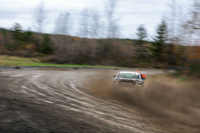 A black car with orange highlights, sliding through a right hand turn. The turn is muddy and muck is being sprayed up by the back tires.