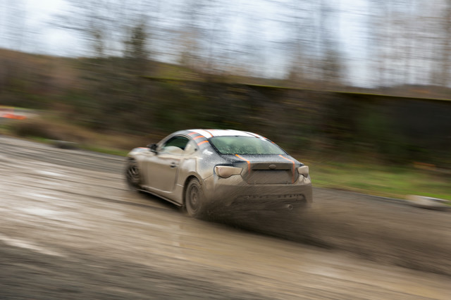 A black car with orange highlights, sliding through a right hand turn. The turn is muddy and muck is being sprayed up by the back tires.