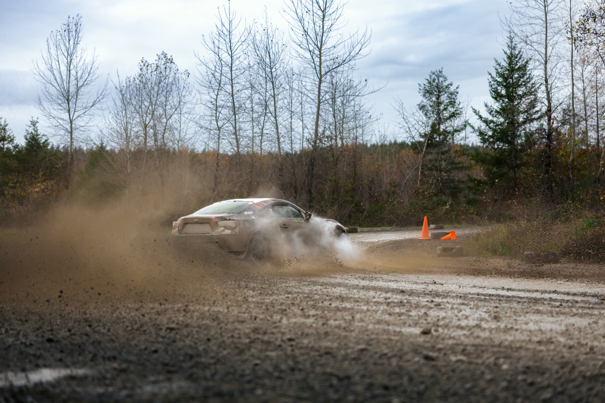 A black car with orange highlights slides through a turn. Muddy spray is kicked up behind it as the front of the car steams from the brakes hitting some water.