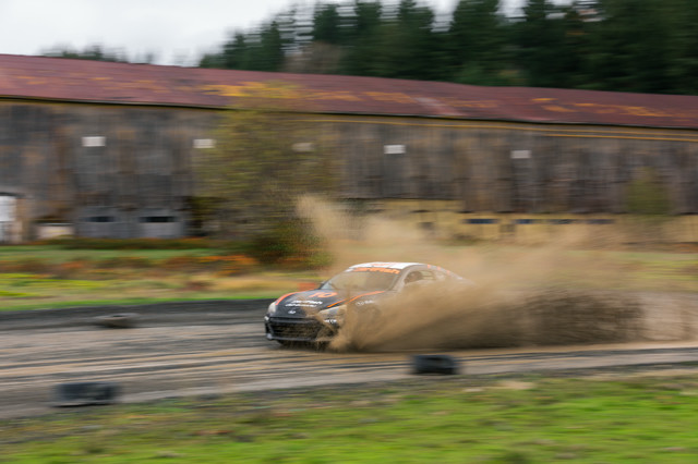 A black car with orange highlights drives through a puddle, creating massive spray. A wooden hanger is in the background.