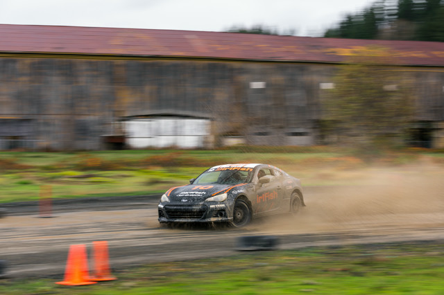 A black car with orange highlights drives through a puddle, creating massive spray. A wooden hanger is in the background.