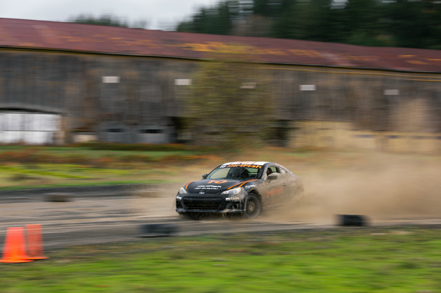 A black car with orange highlights drives through a puddle, creating massive spray. A wooden hanger is in the background. The car is driving sideways with visible counterstreer in the front left wheel.